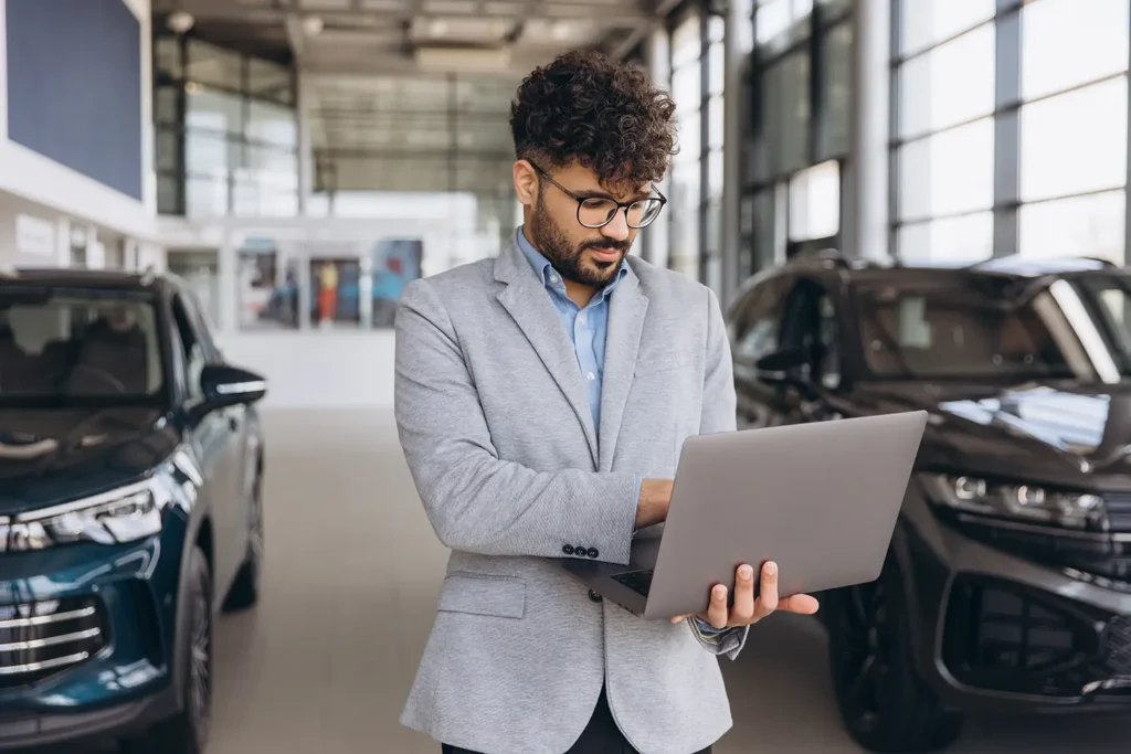 Man using a laptop at a car dealership to perform a dealer invoice price lookup on new vehicles
