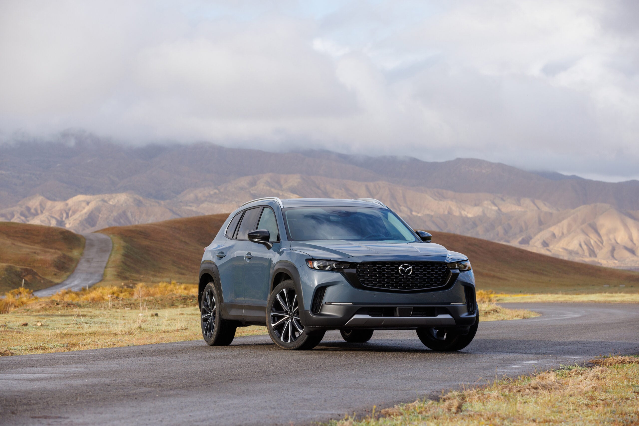 Gray Mazda CX-50 SUV parked on a winding road with mountains and cloudy skies in the background for a Mazda CX-50 Review.