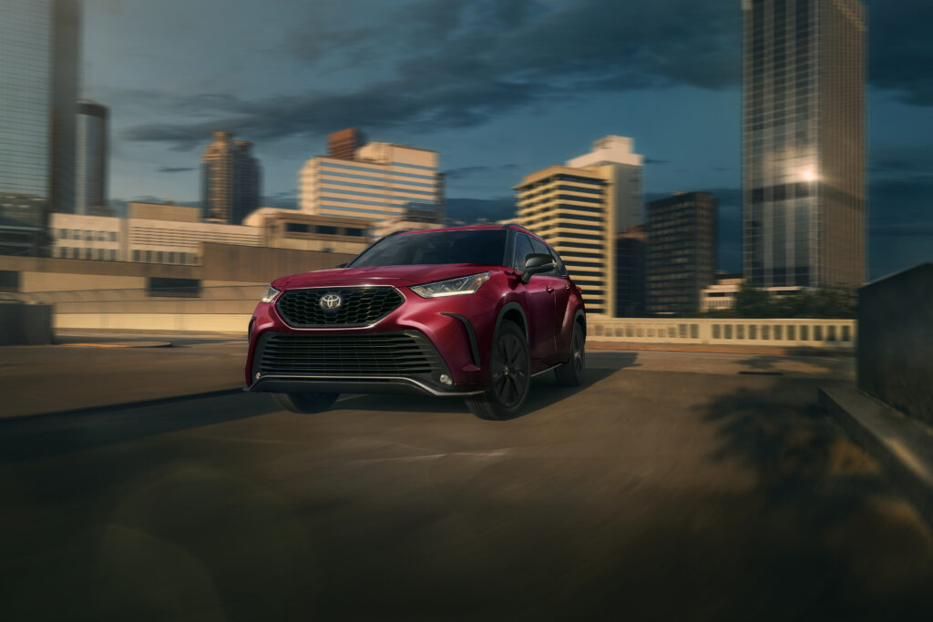 Red Toyota Highlander SUV driving on an urban road with a city skyline in the background for a Toyota Highlander Review.