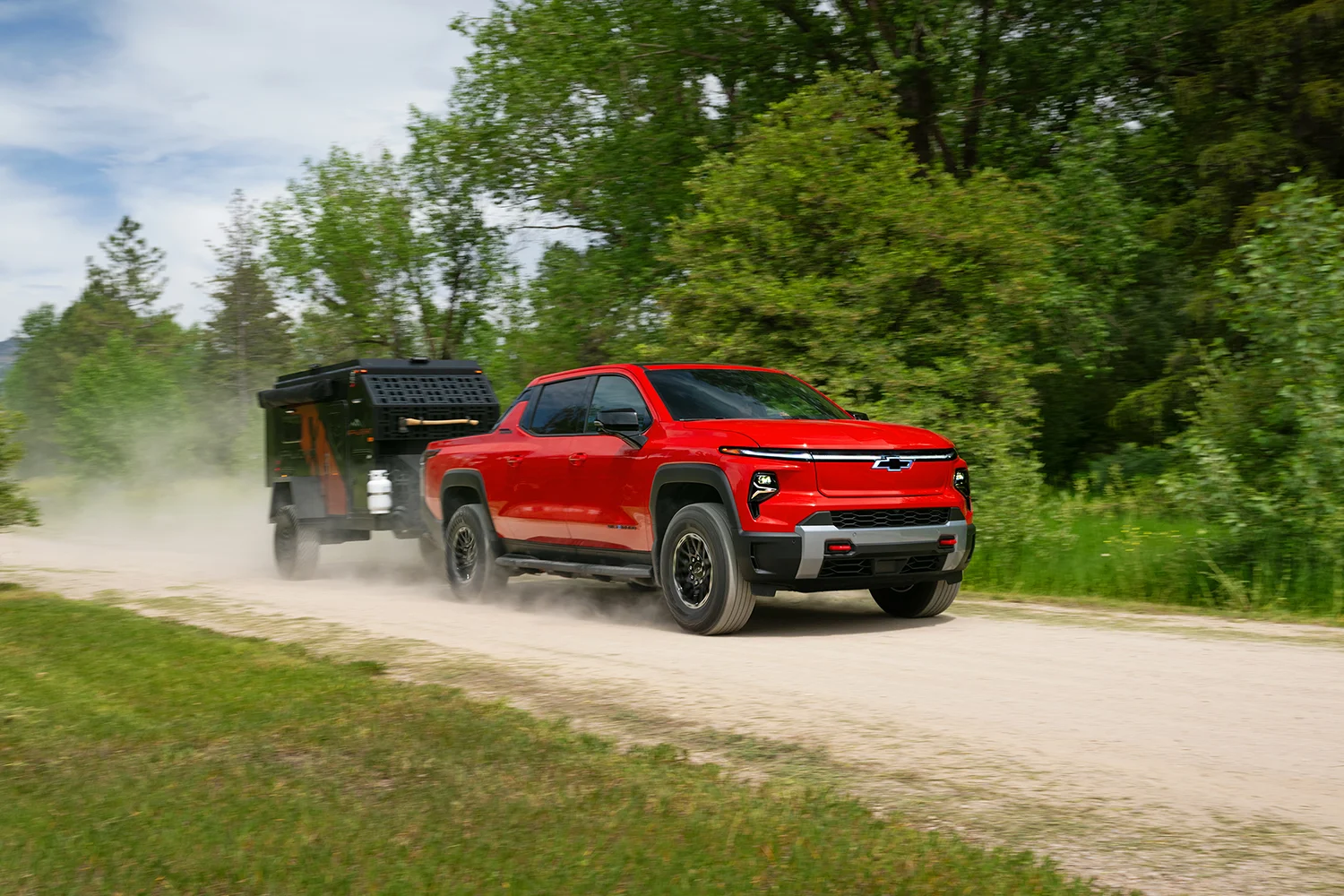 2026 Chevrolet Silverado EV Trail Boss in Radiant Red towing a black off-road camping trailer on a gravel dirt road surrounded by green trees