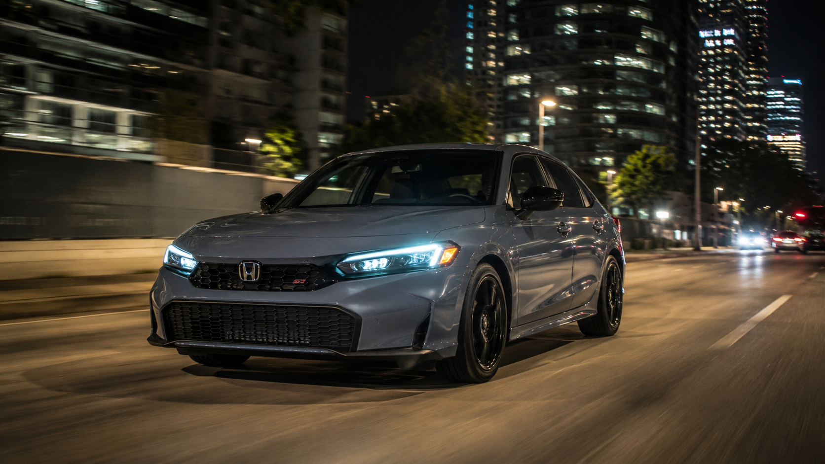2026 Honda Civic Si in Sonic Gray Pearl driving at night through a city with illuminated skyscrapers and street lights reflecting off the bodywork