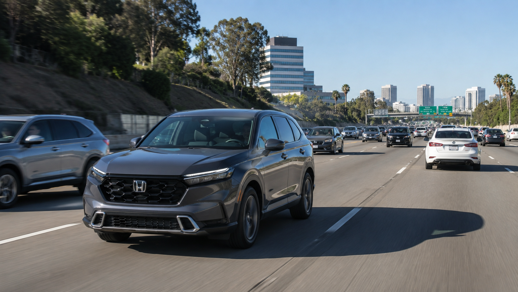 Gray Honda SUV driving in the left lane on a busy highway, with multiple cars ahead and a city skyline in the distance under a clear blue sky