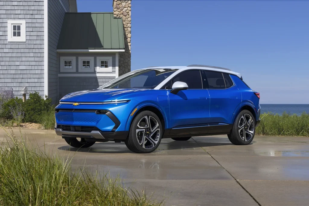2026 Chevrolet Equinox EV in Riptide Blue Metallic with white roof parked on a coastal driveway with ocean and beach grass in the background