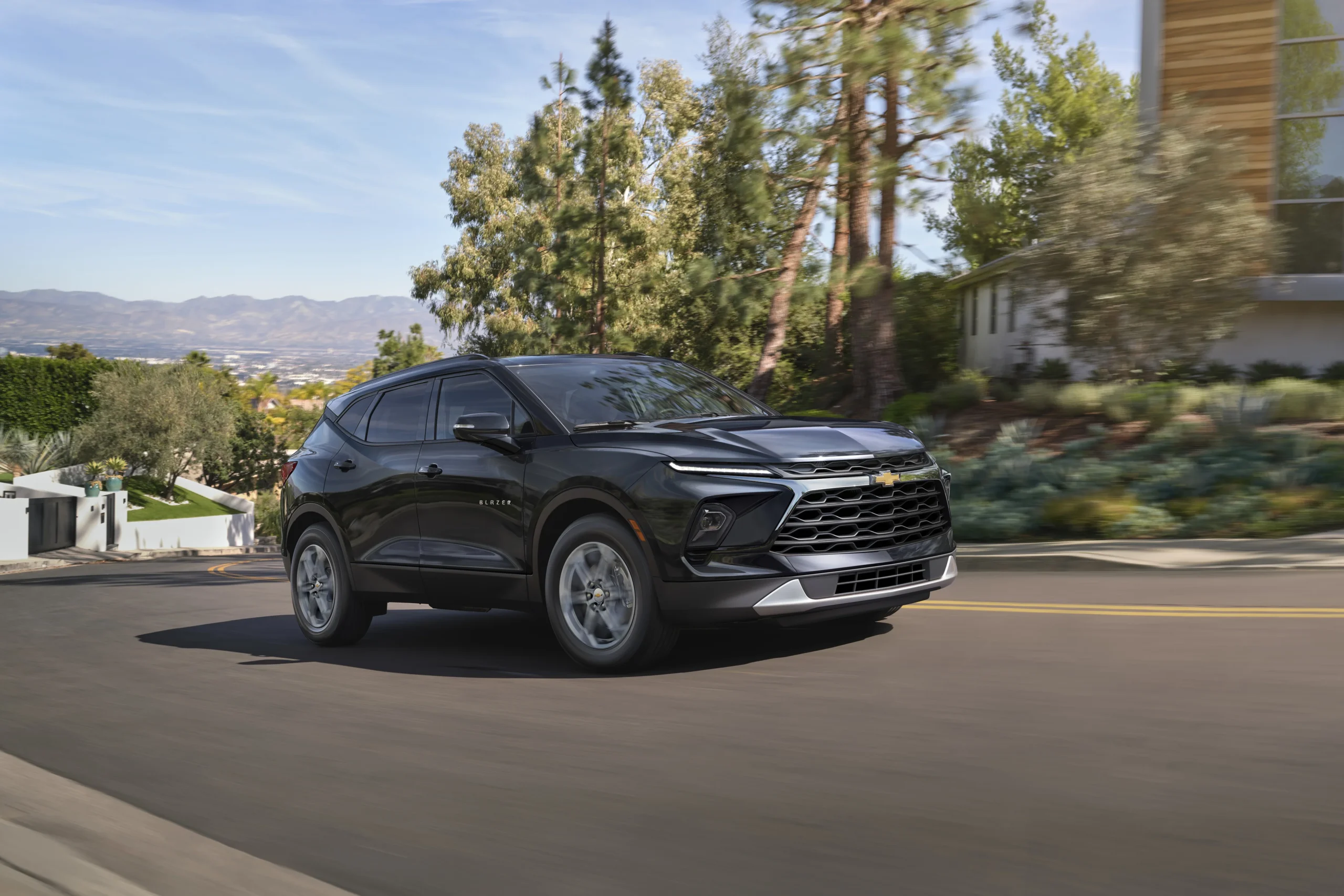 2026 Chevrolet Blazer in black driving on a suburban road with trees, mountains, and modern architecture in the background
