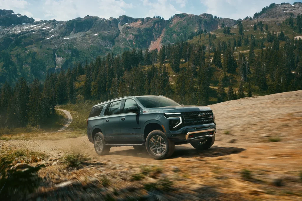 2026 Chevrolet Suburban Z71 in dark teal driving on a dirt mountain trail with snow-capped peaks and pine forest in the background