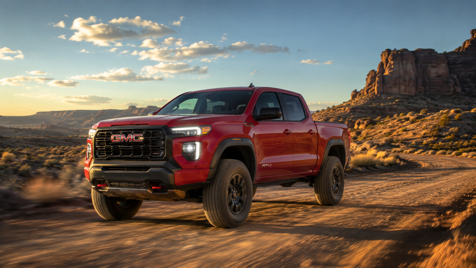 2026 GMC Canyon AT4 in Volcanic Red Tintcoat driving on a desert dirt trail at sunset with red rock formations and a dramatic sky in the background