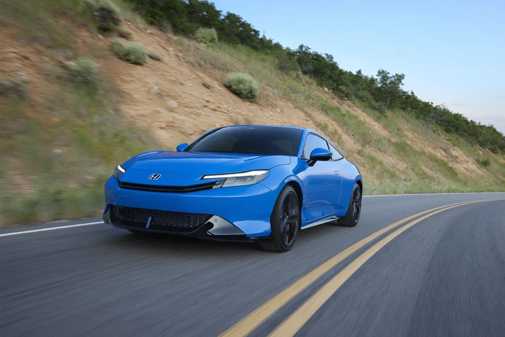 2026 Honda Prelude in Boost Blue Pearl taking a curve on a mountain road at speed with a dry hillside and trees visible in the background