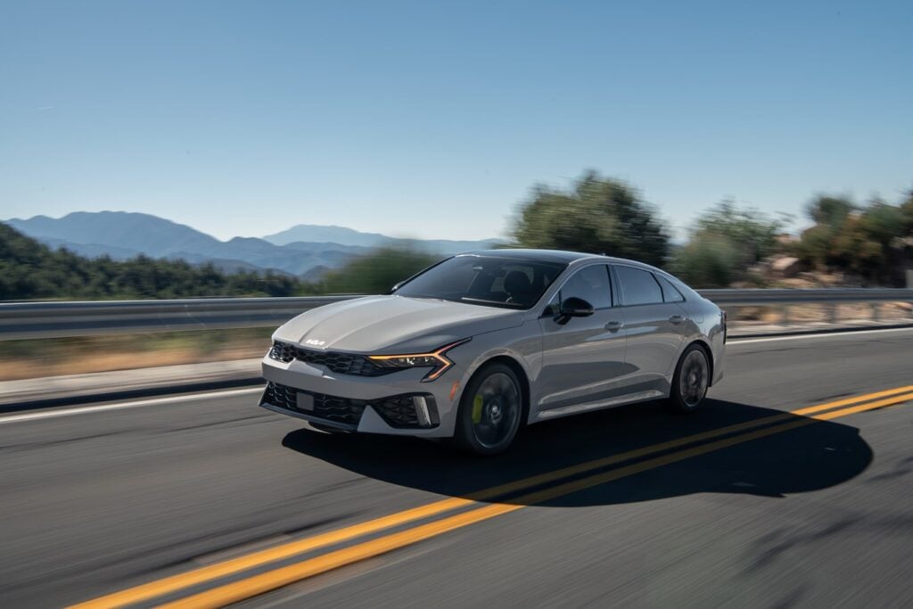 2026 Kia K5 midsize sedan in silver driving on a two-lane road with mountains in the background