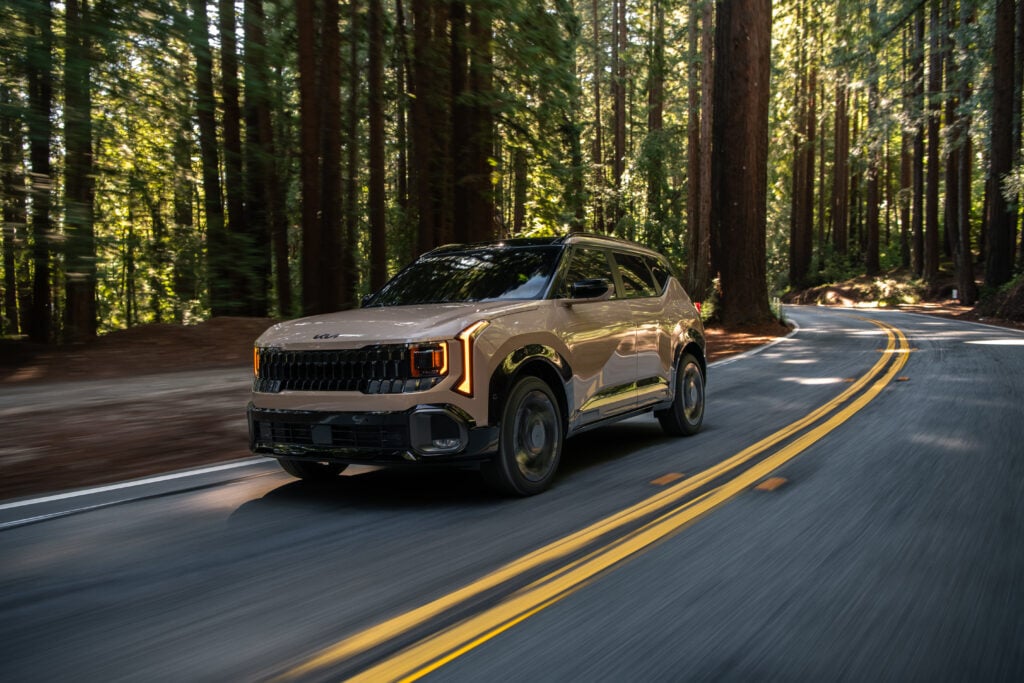 2026 Kia Seltos subcompact SUV in beige driving along a winding forest road with tall trees and double yellow lines