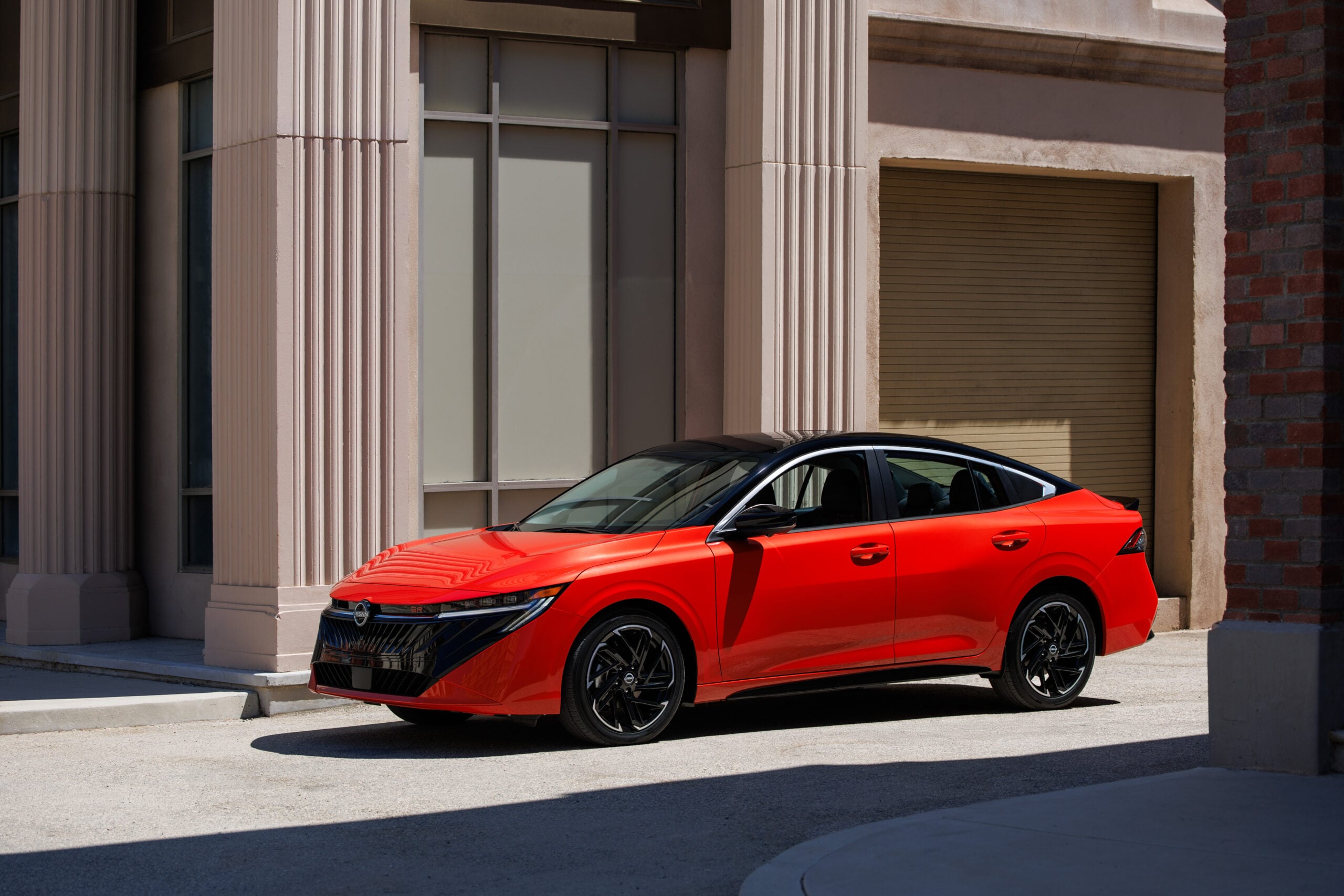 Red 2026 Nissan Sentra sedan parked in front of a beige columned building, showing its redesigned V-motion grille and slim LED headlights.