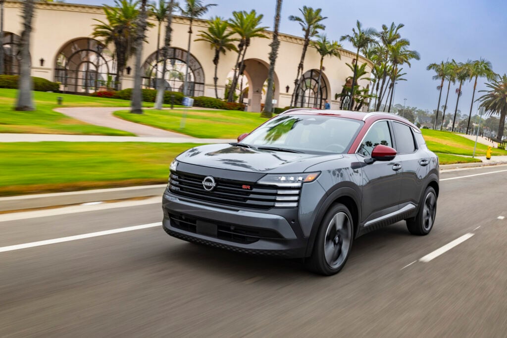 2026 Nissan Kicks SR in two-tone gray and red paint accelerating down a palm tree-lined street past a resort-style building.