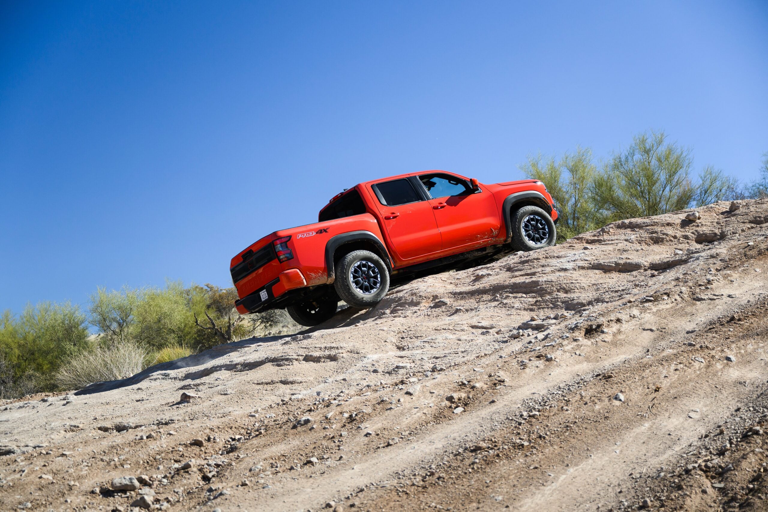Red 2026 Nissan Frontier PRO-4X crew cab pickup truck descending a rocky desert trail under a clear blue sky.