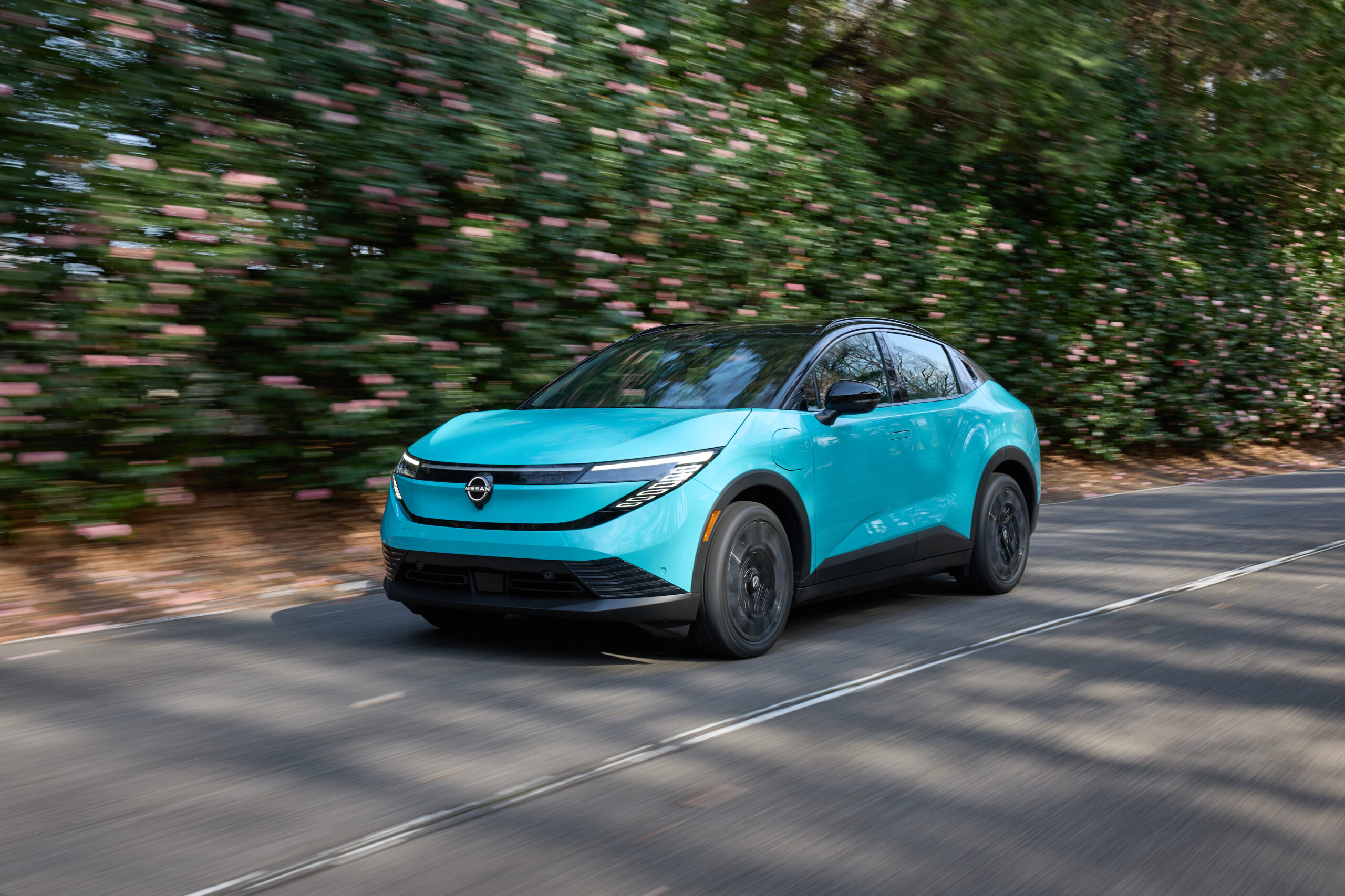 Turquoise 2026 Nissan Leaf driving down a tree-lined road, showing the redesigned crossover-coupe body and closed front fascia.