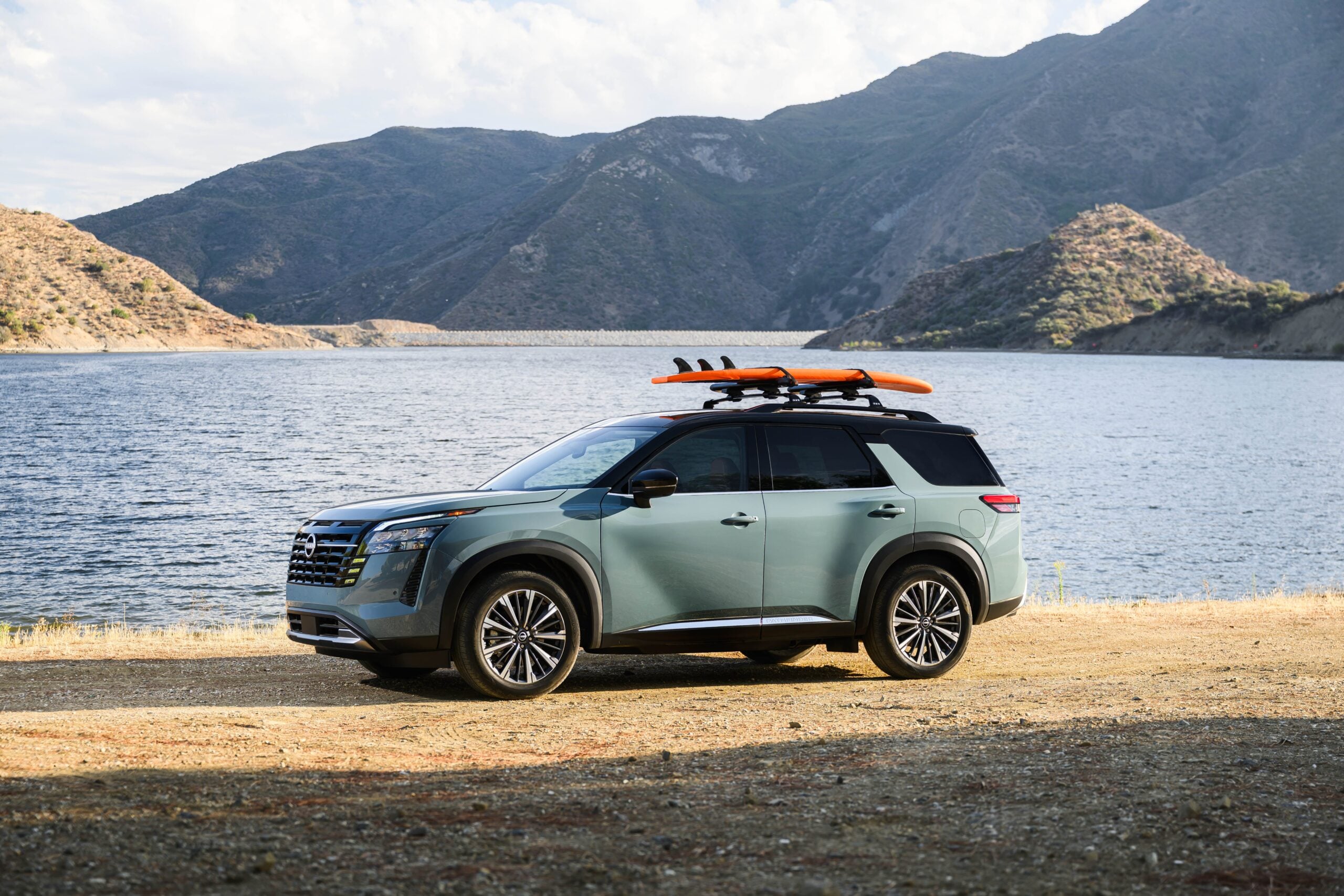 Sage green 2026 Nissan Pathfinder Platinum parked on a rocky lakeshore with a mountain backdrop and an orange surfboard mounted on the roof rack.