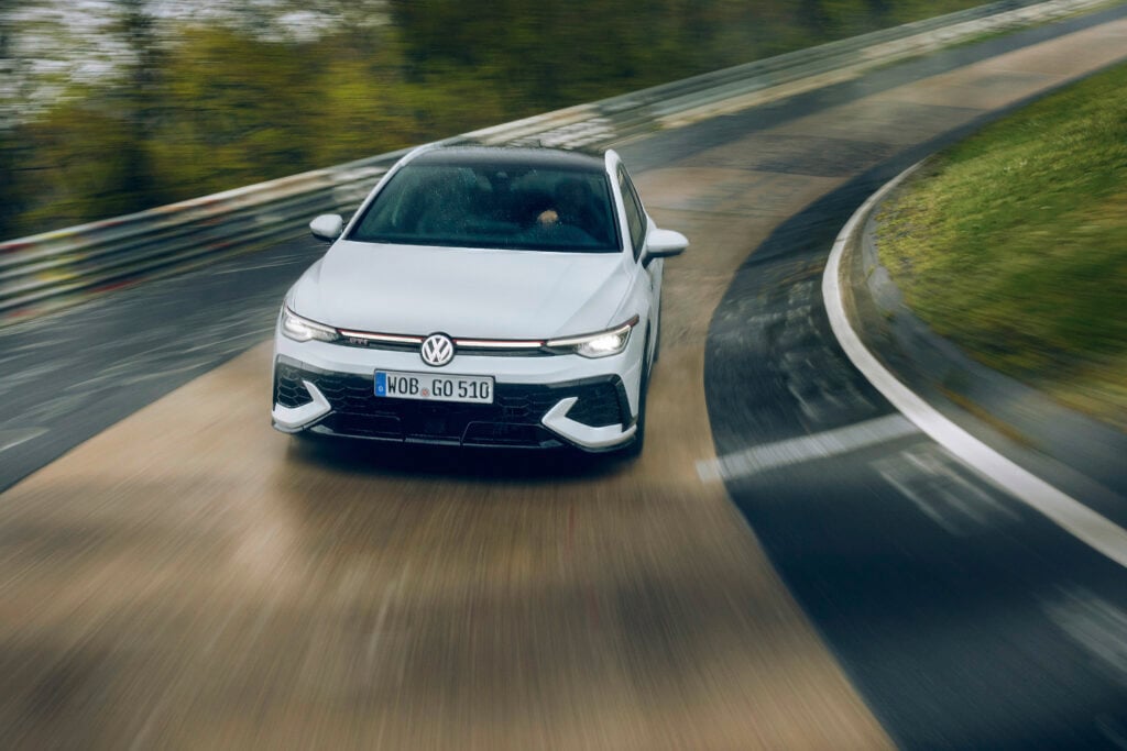 White 2026 Volkswagen Golf GTI with red grille accent driving through a corner on a race track under an overcast sky