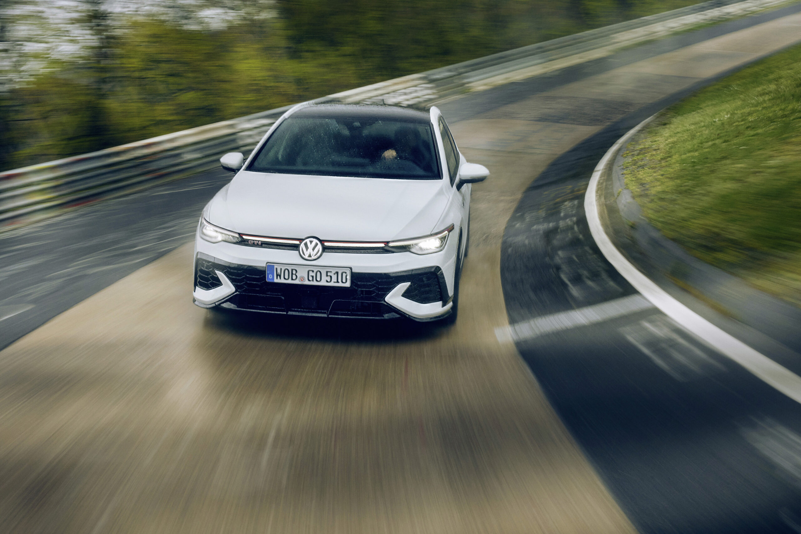 White 2026 Volkswagen Golf GTI with red grille accent driving through a corner on a race track under an overcast sky