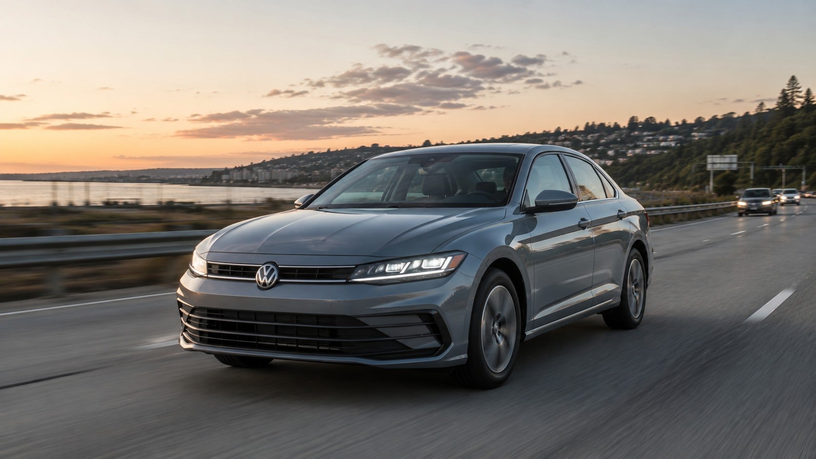 Gray 2026 Volkswagen Jetta sedan driving along a coastal highway at sunset with a hillside town and ocean in the background