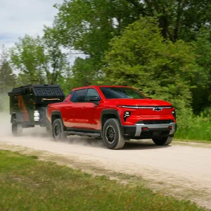 2026 Chevrolet Silverado EV Trail Boss in Radiant Red towing a black off-road camping trailer on a gravel dirt road surrounded by green trees