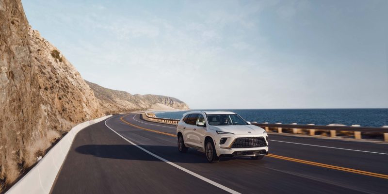 White Buick Enclave driving along a coastal highway beside the ocean, featured in our Buick Enclave Review.