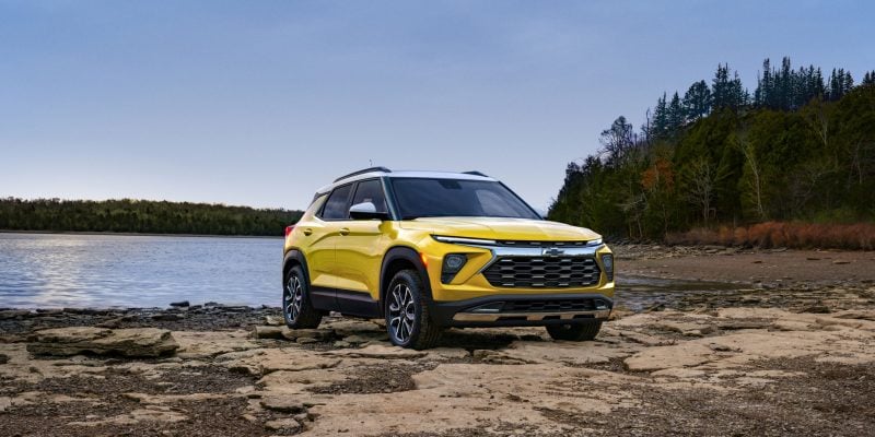 2026 Chevrolet Trailblazer in yellow parked on rocky terrain near a lake with forest in the background