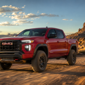 2026 GMC Canyon AT4 in Volcanic Red Tintcoat driving on a desert dirt trail at sunset with red rock formations and a dramatic sky in the background