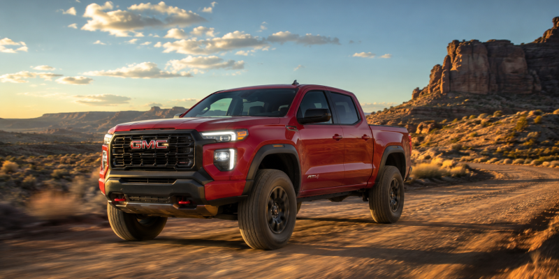 2026 GMC Canyon AT4 in Volcanic Red Tintcoat driving on a desert dirt trail at sunset with red rock formations and a dramatic sky in the background