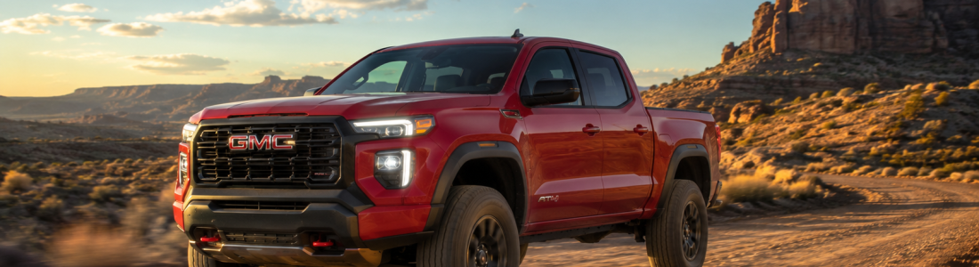 2026 GMC Canyon AT4 in Volcanic Red Tintcoat driving on a desert dirt trail at sunset with red rock formations and a dramatic sky in the background