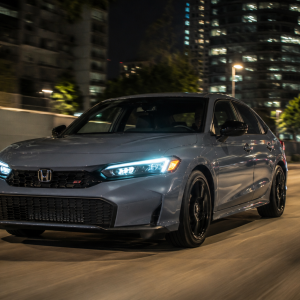2026 Honda Civic Si in Sonic Gray Pearl driving at night through a city with illuminated skyscrapers and street lights reflecting off the bodywork