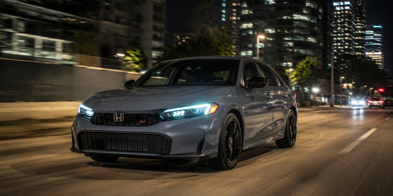 2026 Honda Civic Si in Sonic Gray Pearl driving at night through a city with illuminated skyscrapers and street lights reflecting off the bodywork