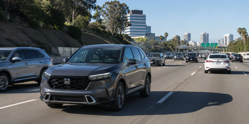 Gray Honda SUV driving in the left lane on a busy highway, with multiple cars ahead and a city skyline in the distance under a clear blue sky