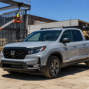 Silver Honda pickup parked on a dusty construction site as two workers in hard hats review blueprints with steel framing and a worker in an elevated lift in the background.