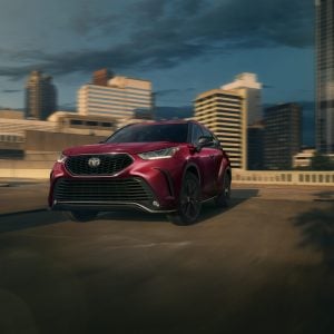 Red Toyota Highlander SUV driving on an urban road with a city skyline in the background for a Toyota Highlander Review.