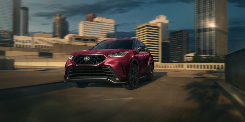 Red Toyota Highlander SUV driving on an urban road with a city skyline in the background for a Toyota Highlander Review.