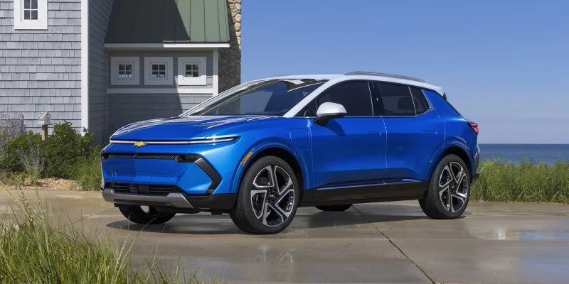 2026 Chevrolet Equinox EV in Riptide Blue Metallic with white roof parked on a coastal driveway with ocean and beach grass in the background