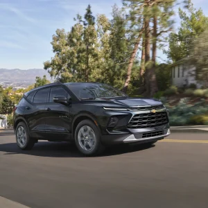 2026 Chevrolet Blazer in black driving on a suburban road with trees, mountains, and modern architecture in the background