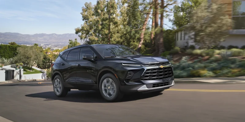2026 Chevrolet Blazer in black driving on a suburban road with trees, mountains, and modern architecture in the background