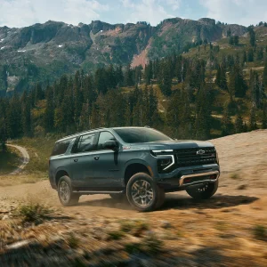 2026 Chevrolet Suburban Z71 in dark teal driving on a dirt mountain trail with snow-capped peaks and pine forest in the background