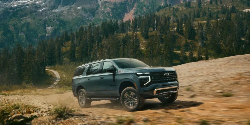 2026 Chevrolet Suburban Z71 in dark teal driving on a dirt mountain trail with snow-capped peaks and pine forest in the background