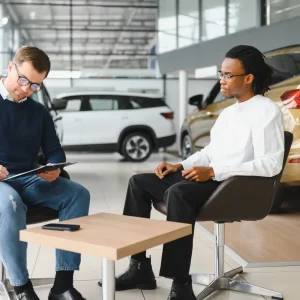 Two men discussing a car invoice at a dealership surrounded by new vehicles.