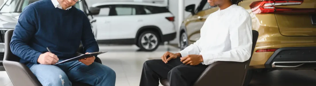 Two men discussing a car invoice at a dealership surrounded by new vehicles.