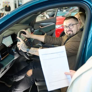 Man sitting in a new car during a dealership visit, reviewing paperwork related to Invoice Price vs. MSRP.