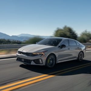2026 Kia K5 midsize sedan in silver driving on a two-lane road with mountains in the background