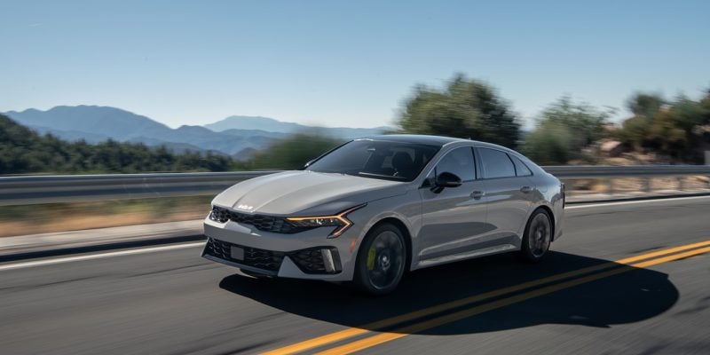 2026 Kia K5 midsize sedan in silver driving on a two-lane road with mountains in the background