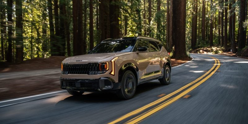 2026 Kia Seltos subcompact SUV in beige driving along a winding forest road with tall trees and double yellow lines