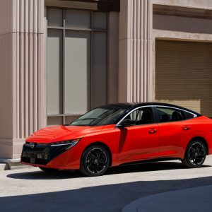 Red 2026 Nissan Sentra sedan parked in front of a beige columned building, showing its redesigned V-motion grille and slim LED headlights.