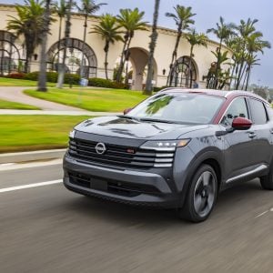 2026 Nissan Kicks SR in two-tone gray and red paint accelerating down a palm tree-lined street past a resort-style building.