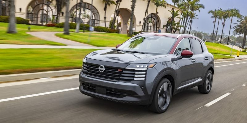 2026 Nissan Kicks SR in two-tone gray and red paint accelerating down a palm tree-lined street past a resort-style building.