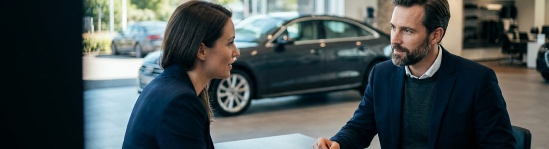 Two professionals in navy blazers discuss paperwork at a desk inside a car showroom, with a sedan visible behind them in the bright space.