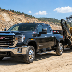2026 GMC Sierra 3500HD in Onyx Black towing a flatbed trailer loaded with a John Deere excavator at a rocky quarry job site under blue skies