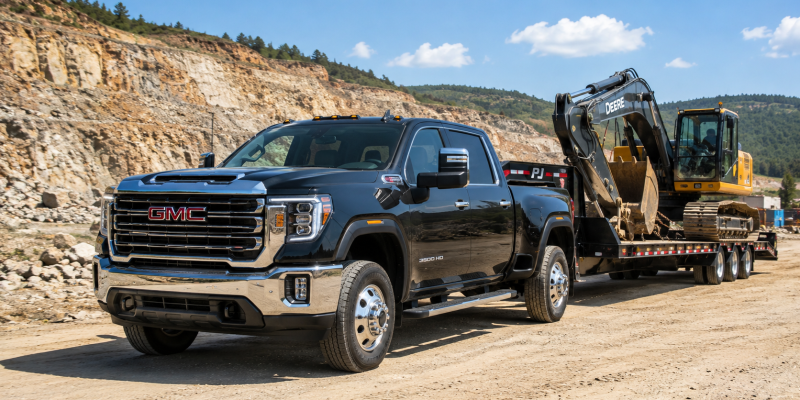 2026 GMC Sierra 3500HD in Onyx Black towing a flatbed trailer loaded with a John Deere excavator at a rocky quarry job site under blue skies